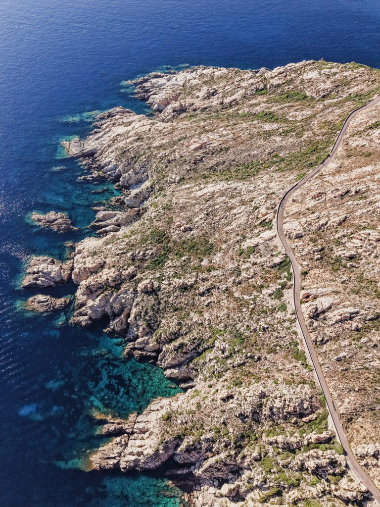 Carretera al borde de un promontorio rocoso sobre el agua turquesa, desde el aire, en Córcega