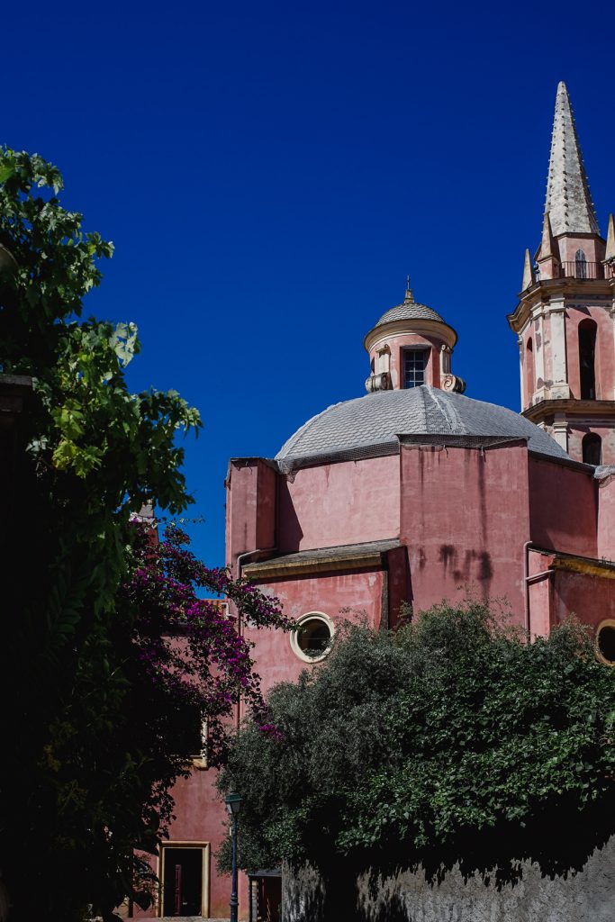Iglesia rosa con cúpula y torre puntiaguda detrás de unos arbustos en Córcega