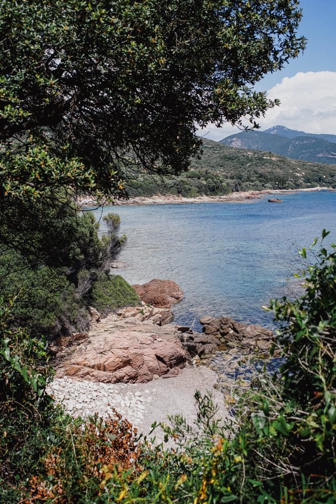 Pequeña cala de guijarros con roca roja, enmarcada por arbustos, en la costa de Córcega