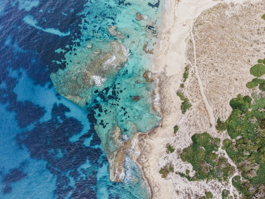 Arena clara y agua turquesa poco profunda con manchas oscuras, desde el aire, en Córcega