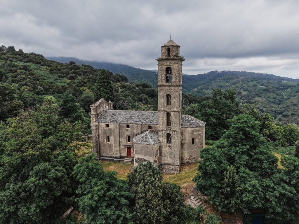 Iglesia de piedra con alto campanario entre colinas verdes, desde el aire, en Córcega