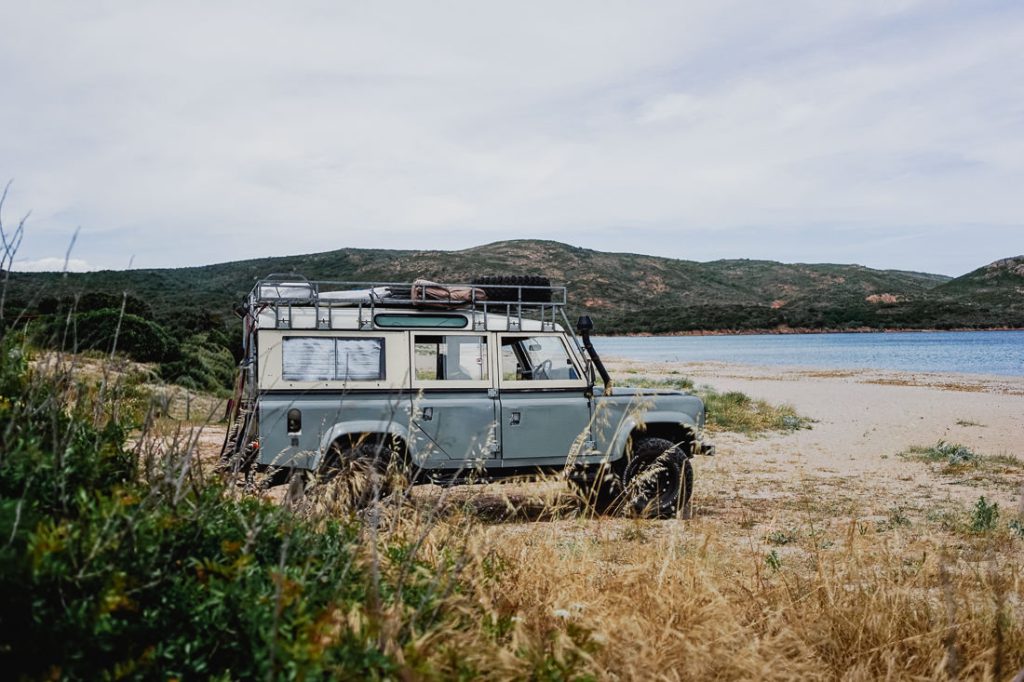Todoterreno gris con baca en la playa, con montañas detrás en Córcega