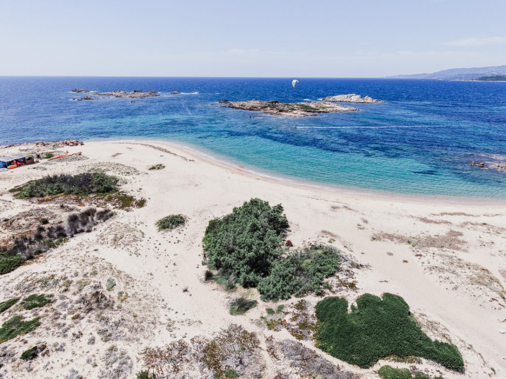 Playa de arena e islotes rocosos, con una vela en el cielo, desde el aire, en Córcega
