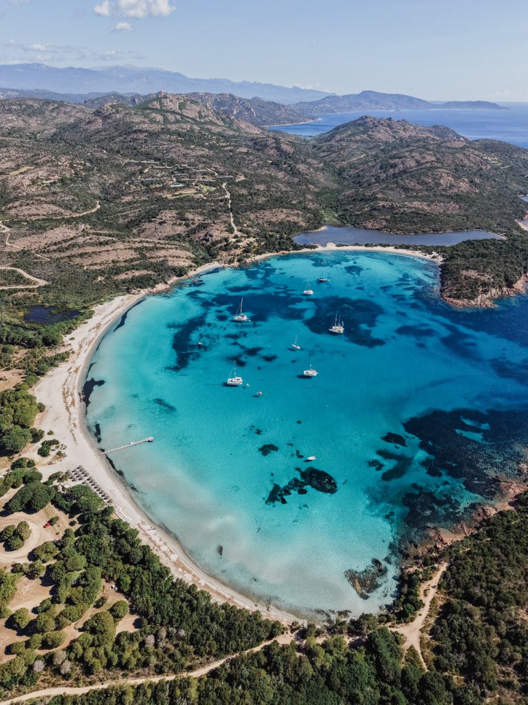 Bahía redonda y turquesa con veleros ante montañas boscosas, desde el aire, en Córcega